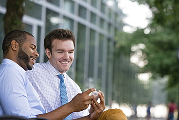 Summer in the city. Businesspeople outdoors, on the go. Two men sitting on a bench, using a smart phone. 