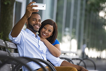 Summer in the city. Businesspeople outdoors, on the go. A couple sitting on a bench, taking a selfy photograph.