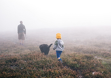 An adult and a child with a dog, walking through heather in autumn mist, England