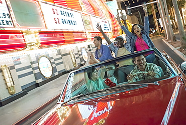 A group of friends in a red open top convertible classic car celebrating with waving arms as they drive through a city lit with neon signs, United States of America