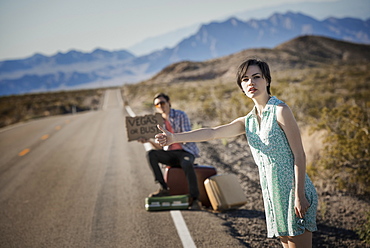 A young couple, man and woman, on a tarmac road in the desert hitchhiking, with a sign saying Vegas or Bust, United States of America