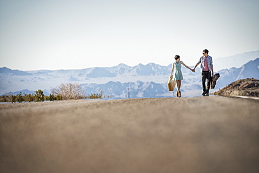 A young couple, man and woman walking hand in hand on a tarmac road in the desert carrying cases, United States of America