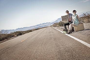 A young couple, man and woman, on a tarmac road in the desert hitchhiking, with a sign saying Vegas or Bust, United States of America