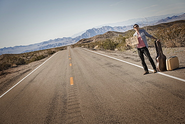 A man standing by the roadside, a hitchhiker with guitar and case, holding a sign saying Vegas or Bust, United States of America