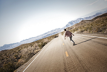 A young man riding down a tarmac road in the desert on a skateboard, United States of America