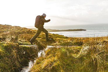 A man with a rucksack and winter clothing leaping across a small stream in an open exposed landscape, Scotland, United Kingdom