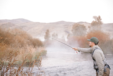 A man standing casting his fishing rod, flyfishing from a riverbank.