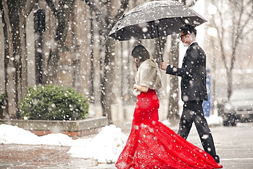 A woman in a long red evening dress with fishtail skirt wearing a fur stole, and a man in a suit, walking through snow in the city.