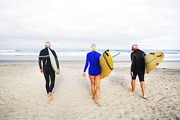 Senior woman and two senior men on a beach, wearing wetsuits and carrying surfboards, United States of America