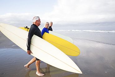 Senior woman and two senior men on a beach, wearing wetsuits and carrying surfboards, United States of America