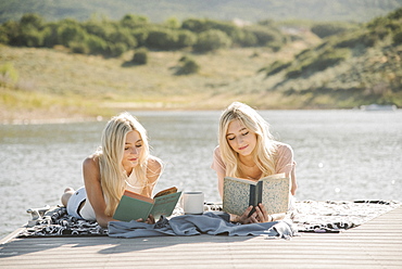 Two blond sisters lying on a jetty, reading a book.