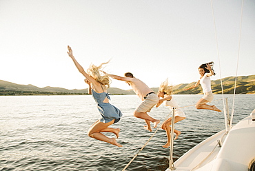 Four people jumping off the side of a sail boat into a lake.