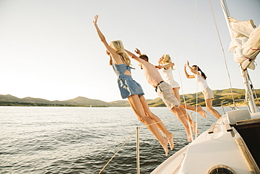 Four people jumping off the side of a sail boat into a lake.