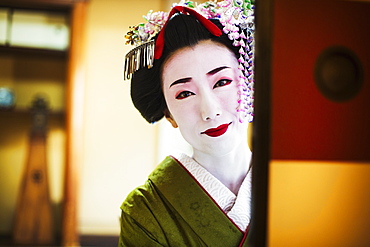 A woman dressed in the traditional geisha style, wearing a kimono and obi, with an elaborate hairstyle and floral hair clips, with white face makeup with bright red lips and dark eyes, looking in the mirror, Japan