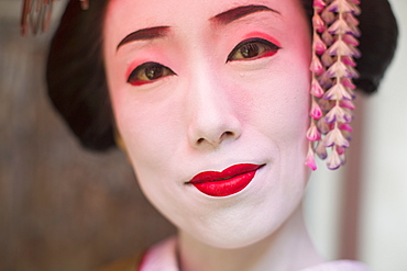 A woman made up in traditional geisha style with an elaborate hairstyle and floral hair clips, drawn eyebrows with white face makeup with bright red lips and outlined eyes, Japan