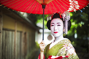 A woman dressed in the traditional geisha style, wearing a kimono and obi, with an elaborate hairstyle and floral hair clips, with white face makeup with bright red lips and dark eyes holding a red paper parasol, Japan