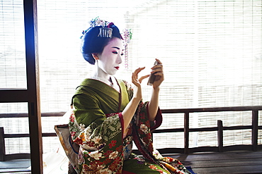 A woman dressed in the traditional geisha style, wearing a kimono and obi, with an elaborate hairstyle and floral hair clips, with white face makeup with bright red lips and dark eyes taking a selfie of herself, Japan