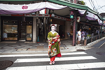 A woman dressed in the traditional geisha style, wearing a kimono and obi, with an elaborate hairstyle and floral hair clips, with white face makeup with bright red lips and dark eyes crossing a street, Japan