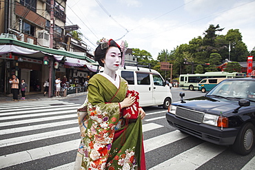 A woman dressed in the traditional geisha style, wearing a kimono and obi, with an elaborate hairstyle and floral hair clips, with white face makeup with bright red lips and dark eyes crossing a street, Japan