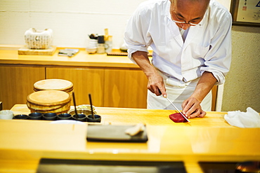 A chef working in a small commercial kitchen, an itamae or master chef slicing fish with a large knife for making sushi, Japan