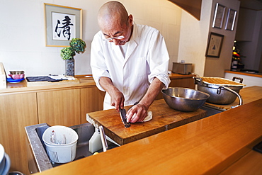 A chef working in a small commercial kitchen, an itamae or master chef slicing fish with a large knife for making sushi, Japan