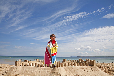 A boy standing beside a sandcastle, on top of a mound of sand. Beach, England