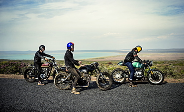 Three men wearing open face crash helmets sitting on cafe racer motorcycles on a rural road, United States of America