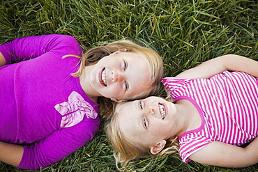 A portrait of two sisters smiling. View from above, of two girls lying on their backs on the grass, heads together.