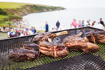 A beach barbecue with meat cooking over an open fire. People in the background on the cliffs overlooking the coastline, England, United Kingdom