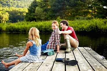 Three people, two adults and a child relaxing on a jetty, with their feet in the water at the end of a day, Kingston, New York, USA