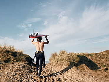 Man walking on a path in the sand dunes towards sea with surf board balancing on his head, United Kingdom