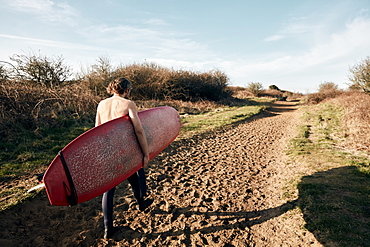 Man holding surf board walking along sandy path towards beach, United Kingdom