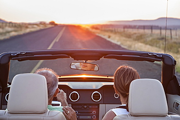 View from behind of senior couple in a convertible sports car driving on a highway at sunset in eastern Washington State, United States of America