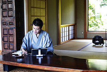 Japanese man wearing kimono sitting on floor in traditional Japanese house, looking at digital tablet, Kyushu, Japan