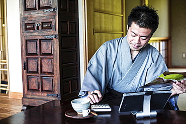 Japanese man wearing kimono sitting on floor in traditional Japanese house, using calculator and digital tablet, Kyushu, Japan