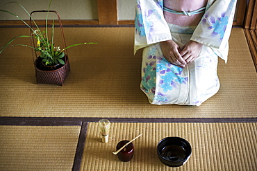 Japanese woman wearing traditional white kimono with blue floral pattern kneeling on tatami mat in front of bowl and whisk for tea ceremony, Kyushu, Japan