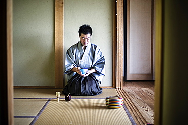 Japanese man wearing traditional kimono kneeling on tatami mat, holding tea bowl, during tea ceremony, Kyushu, Japan