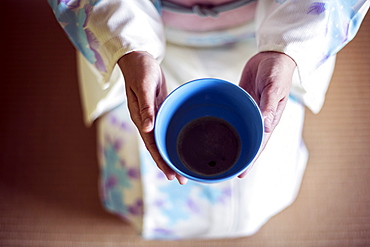 High angle close up of Japanese woman wearing traditional white kimono with blue floral pattern kneeling on floor during tea ceremony, holding blue tea bowl, Kyushu, Japan