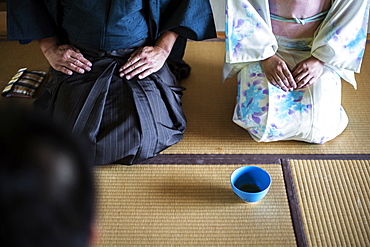 High angle view of Japanese man and woman wearing traditional white kimono with blue floral pattern kneeling on floor during tea ceremony, holding blue tea bowl, Kyushu, Japan