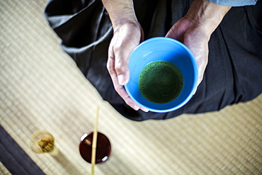 High angle close up of Japanese man wearing traditional kimono kneeling on floor holding blue bowl with Matcha tea during tea ceremony, Kyushu, Japan