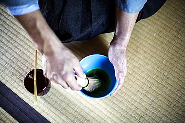 High angle close up of Japanese man wearing traditional kimono kneeling on floor using bamboo whisk to prepare Matcha tea in a blue bowl during tea ceremony, Kyushu, Japan