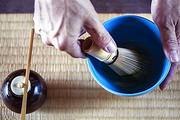 High angle close up of person using bamboo whisk to prepare Matcha tea in a blue bowl during tea ceremony, Kyushu, Japan