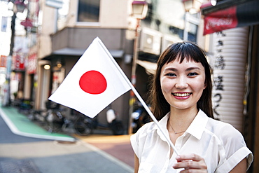 Smiling Japanese woman with long brown hair wearing white short-sleeved blouse standing in a street, holding small Japanese flag, Kyushu, Japan