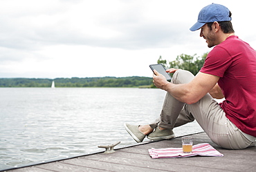 A man seated on a jetty by a lake, using a digital tablet.
