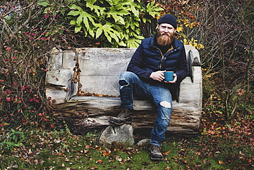 Bearded man wearing black beanie sitting on wooden bench in garden, holding blue mug, looking at camera, Berkshire, England