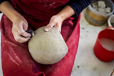 High angle close up of ceramic artist wearing red apron sitting in her workshop, working on clay vase, England