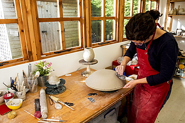 Woman wearing red apron standing in her ceramics workshop, working on clay bowl, England