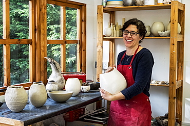 Woman wearing red apron standing in her workshop, holding ceramic vase, smiling at camera, England