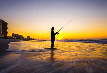 Blurred view of silhouette of man fishing in waves on beach at sunset