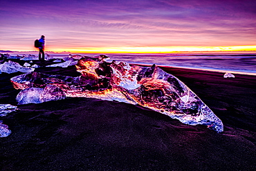 Hiker admiring glaciers on beach reflecting sunset sky, Jokulsarlon, Iceland